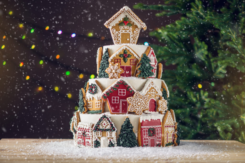 Large tiered Christmas cake decorated with gingerbread cookies and a house on top. Tree and garlands in the background.