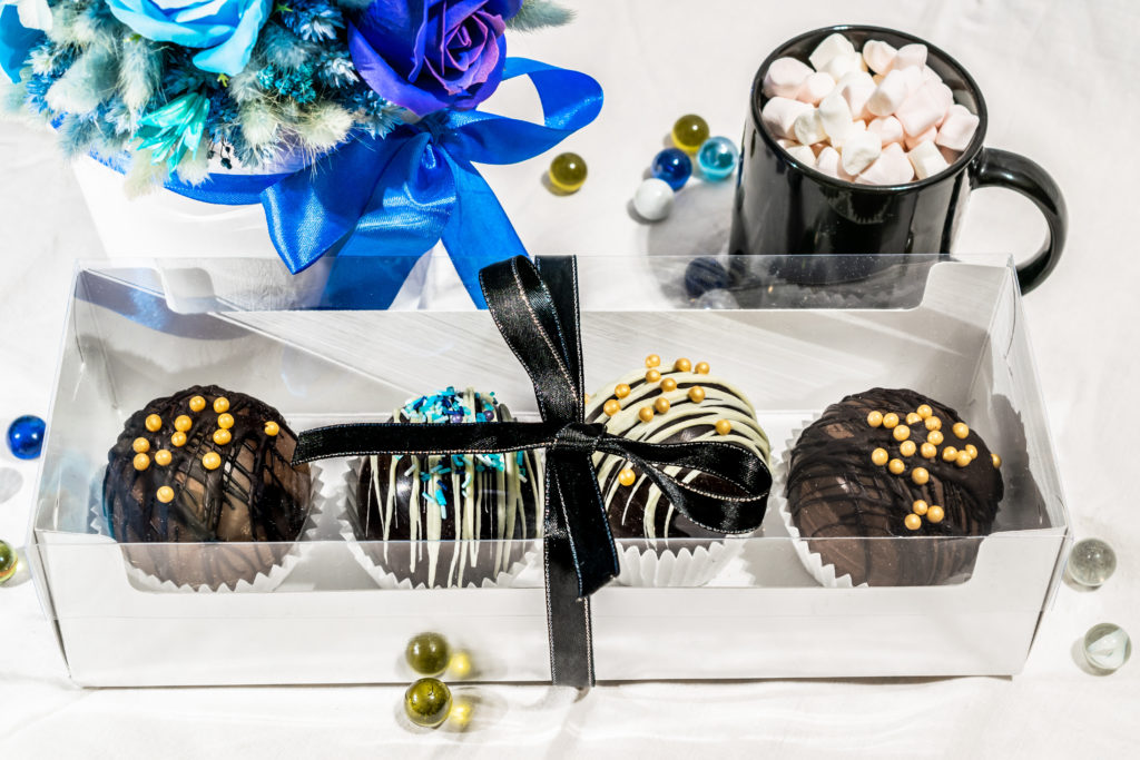 Set of chocolate bombs in giftbox with mini marshmallows, white chocolate, milk chocolate, hot cocoa, ingredients that melts with hot milk. Delicious winter holidays dessert with bouquet of blue roses