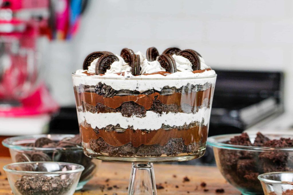 Picture of an oreo brownie trifle in a tall glass dish, layers of brownie, chocolate ganache, whipped cream and decorated with pipped whipped cream and mini oreos. Table also shows bowls of ingredients, such as brownies, whole oreos and crushed oreos