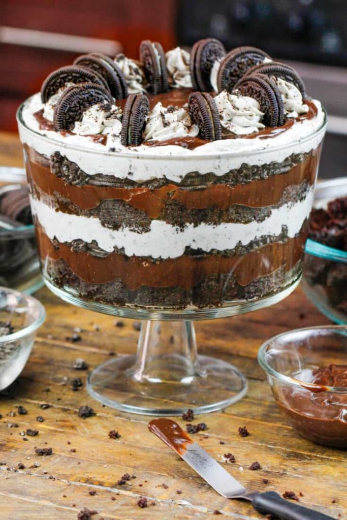 Picture of an oreo brownie trifle in a tall glass dish, layers of brownie, chocolate ganache, whipped cream and decorated with pipped whipped cream and mini oreos