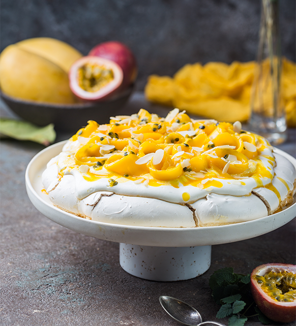 Passionfruit pavlova with whipped cream and fresh passionfruit pulp. In the background there is a mango and sliced in half passionfruit sitting in a black bowl. Bottom right corner, a teaspoon, leaf and half cut passionfruit showing the pulp.